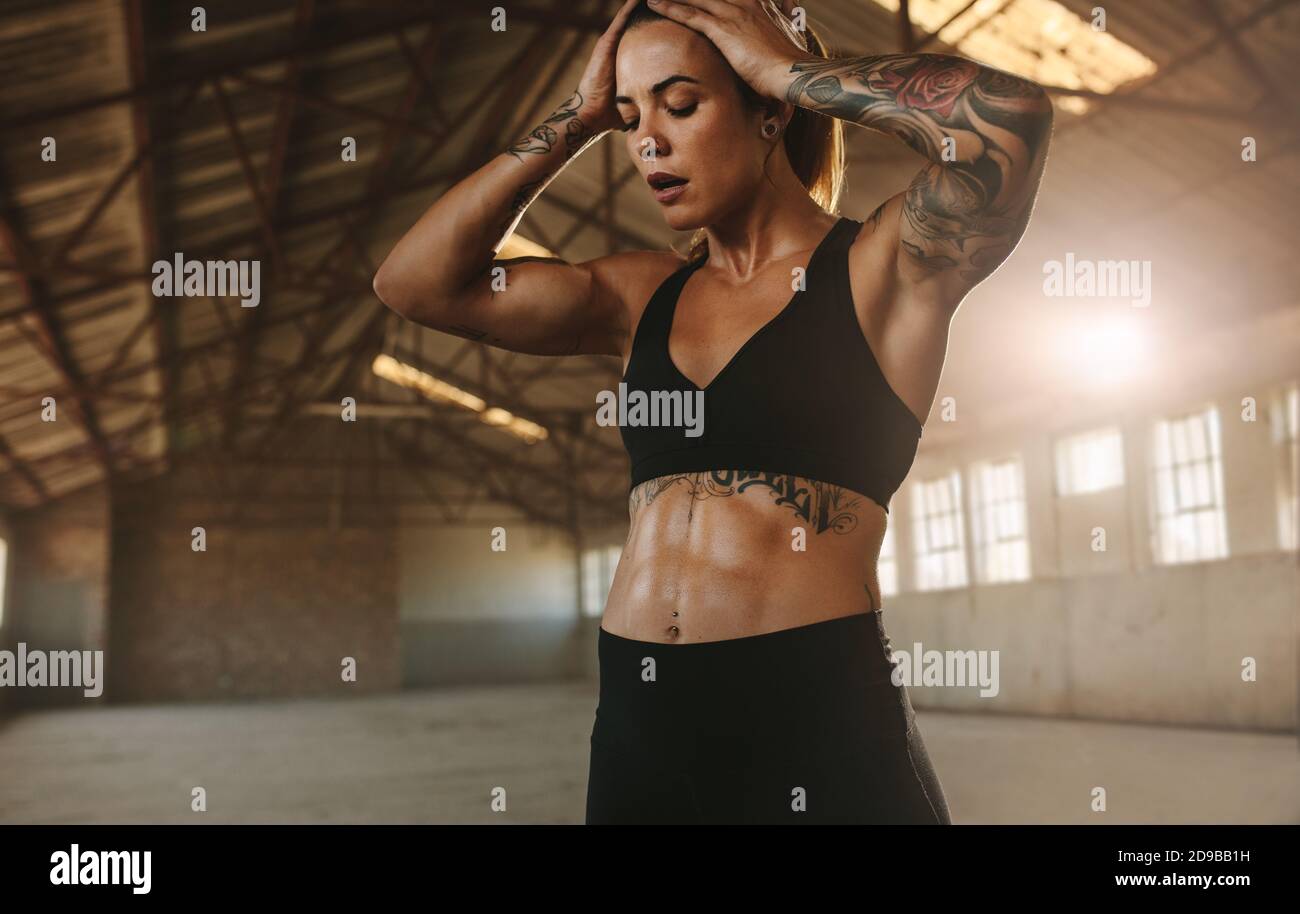 Woman feeling exhausted after intense workout. Female in sports wear standing with her hand on head inside cross training space in old warehouse. Stock Photo