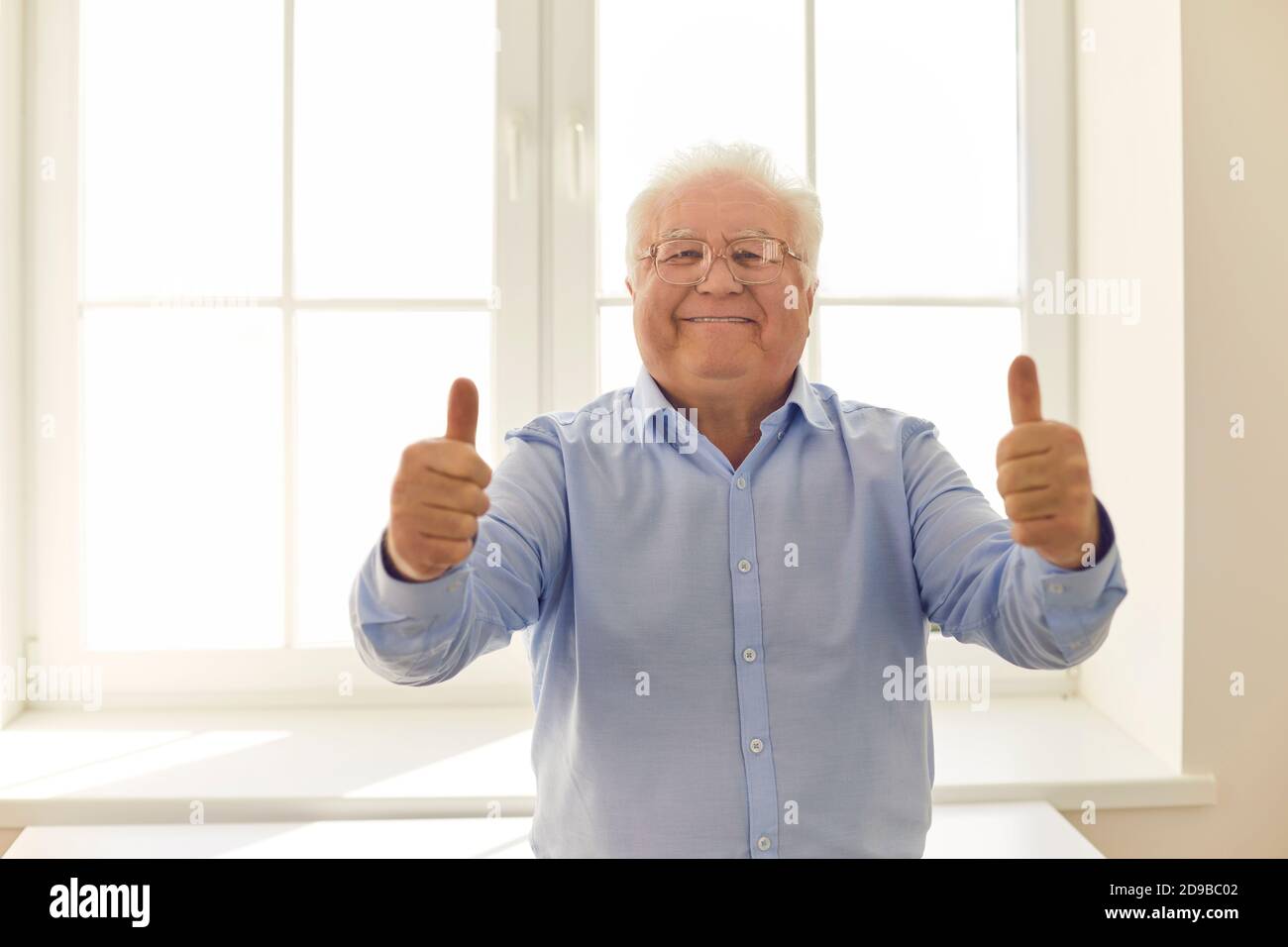 Happy senior man giving thumbs-up and looking at camera standing near the window Stock Photo