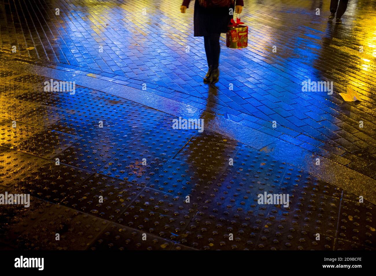 A pedestrian carries a boxed panetone Italian Christmas cake as she crosses the brick paved road illuminated with neon signs after a rain sower had left the roads wet outside the Old Vic Theatre near Waterloo Station in London as the Christmas season is in full-swing. 17 December 2015. Photo: Neil Turner Stock Photo