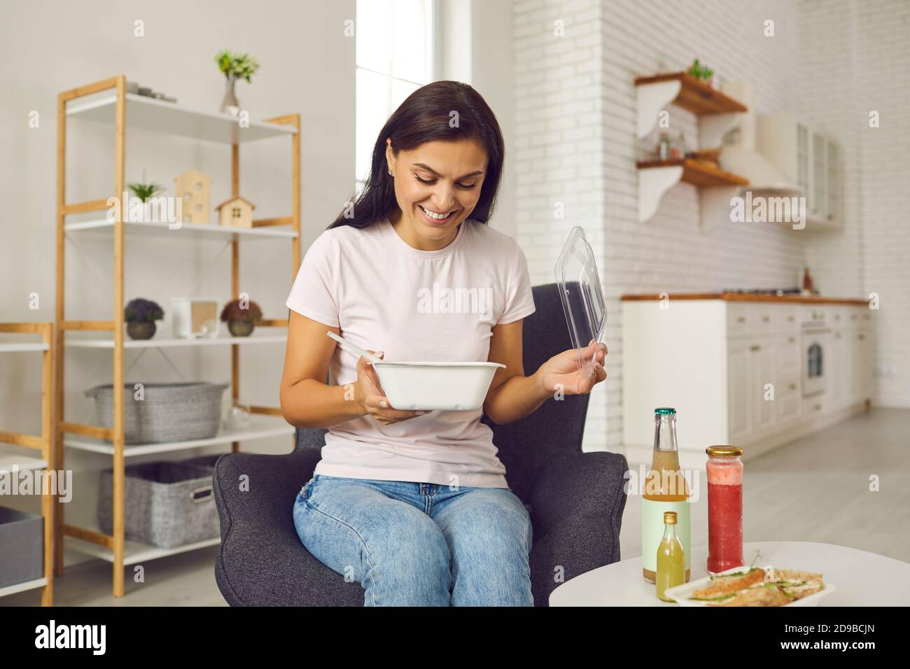 Happy woman sitting at home on and eating takeaway food from containers during lunch break. Stock Photo