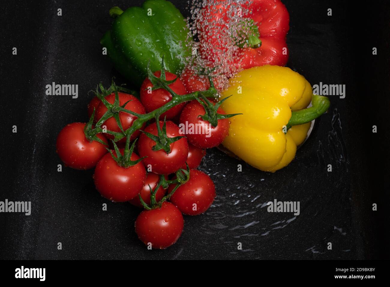 Fresh bell peppers and tomatoes on vine getting washed under tap water in black sink as healthy eating preparing vegetables concept Stock Photo