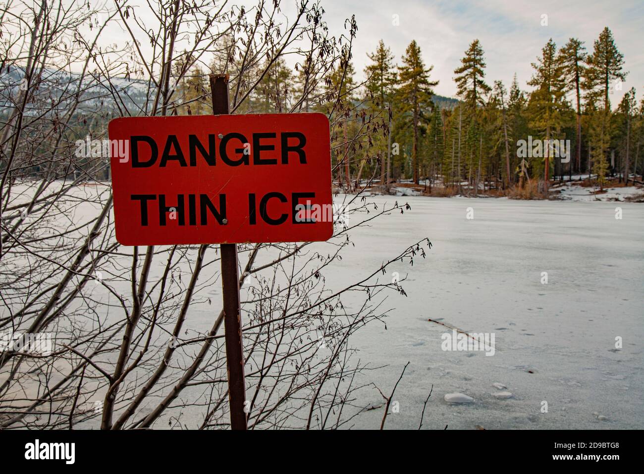 Thin ice sign indicating danger/hazard on frozen over mountain lake (Manzanita Lake in Lassen Volcanic National Park). Stock Photo