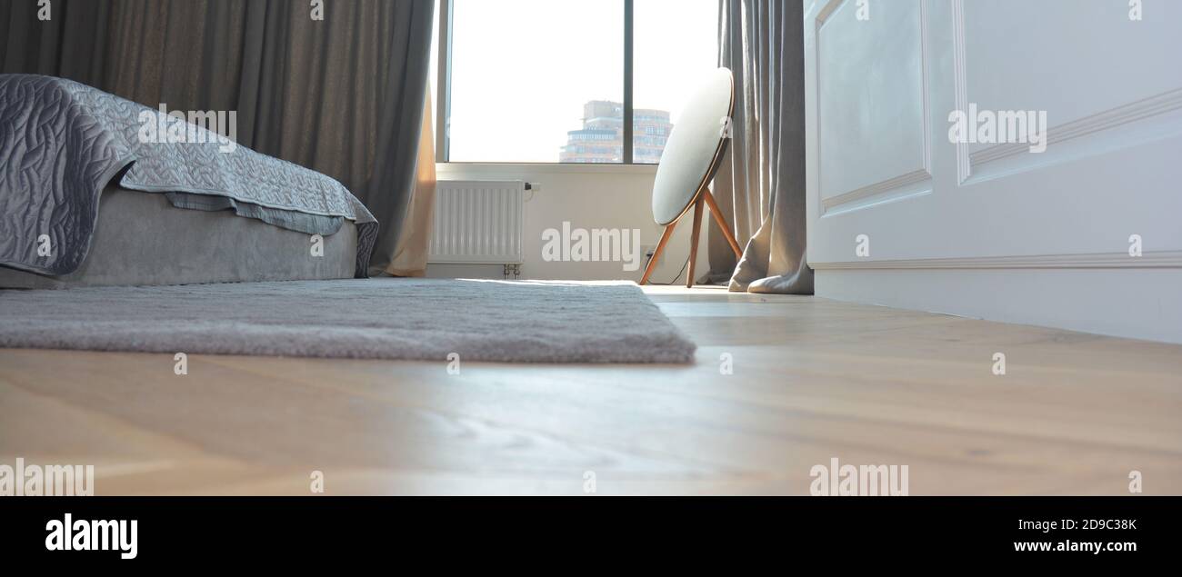 A cosy bedroom with wooden laminated flooring, gray soft carpet, a bed, dark gray curtains on a window and white wood paneled wall. Stock Photo