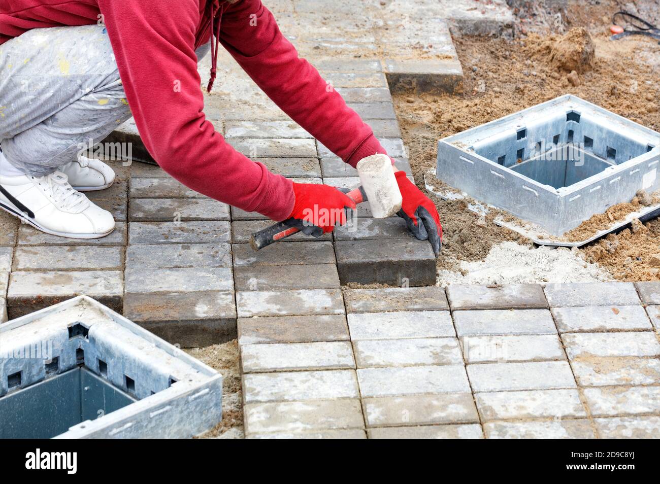 A worker lays down paving slabs and places them on prepared flat sandy soil on the sidewalk around utility hatches, copy space.. Stock Photo