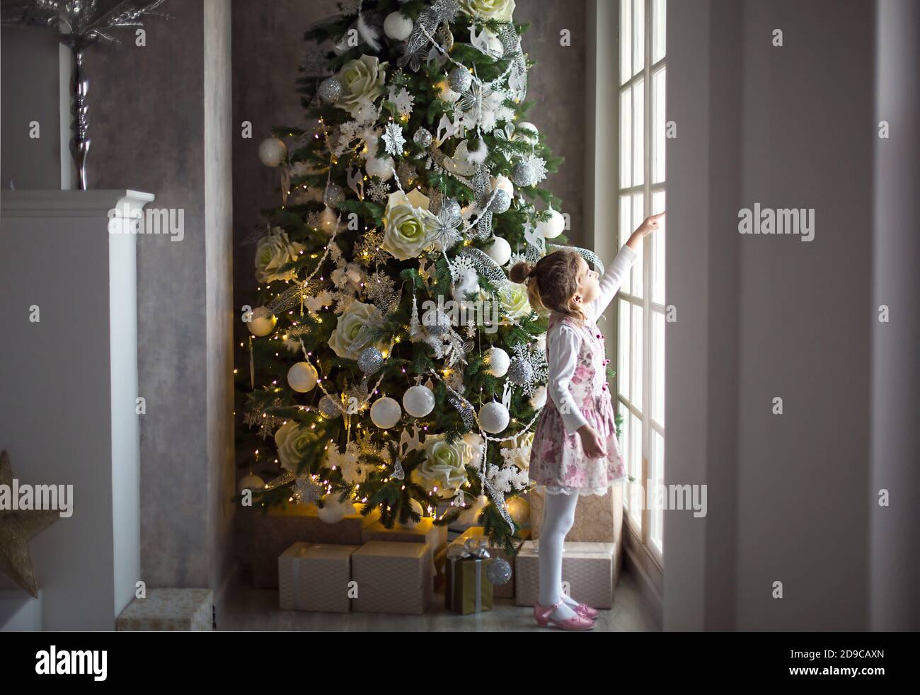 A little girl looks out of a large window near a Christmas tree. Waiting for a miracle, Christmas white decor in the living room of the house. New yea Stock Photo