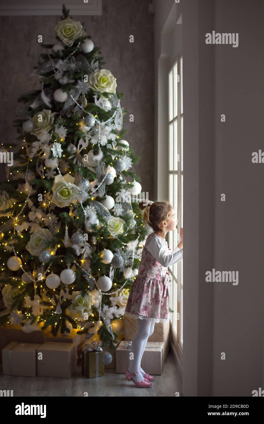 A little girl looks out of a large window near a Christmas tree. Waiting for a miracle, Christmas white decor in the living room of the house. New yea Stock Photo