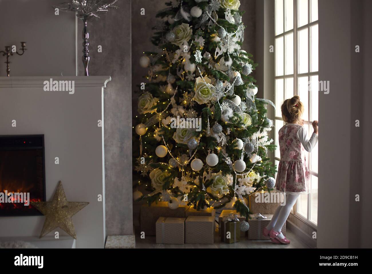 A little girl looks out of a large window near a Christmas tree. Waiting for a miracle, Christmas white decor in the living room of the house. New yea Stock Photo