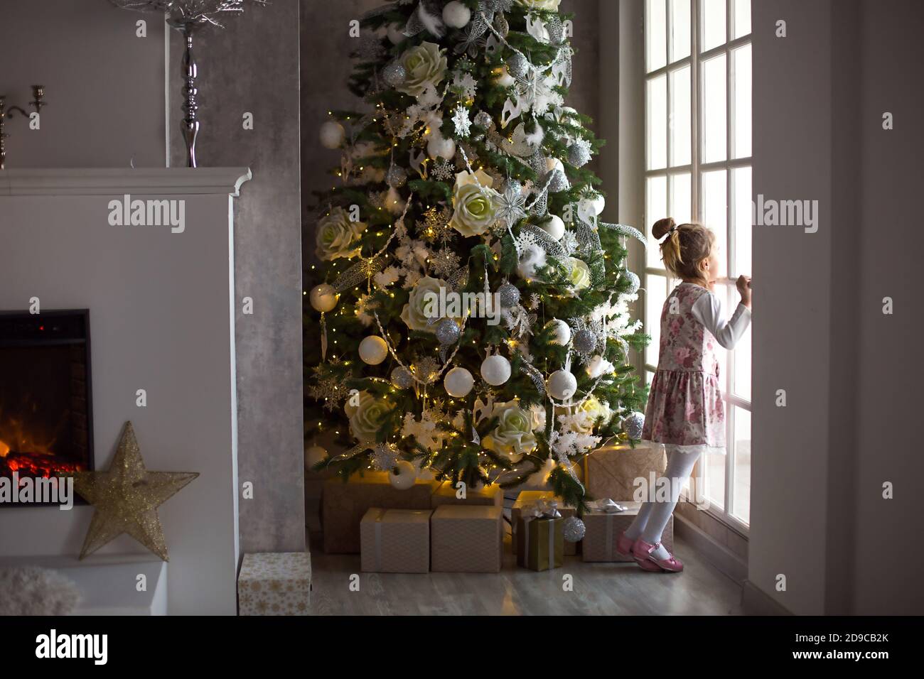 A little girl looks out of a large window near a Christmas tree. Waiting for a miracle, Christmas white decor in the living room of the house. New yea Stock Photo