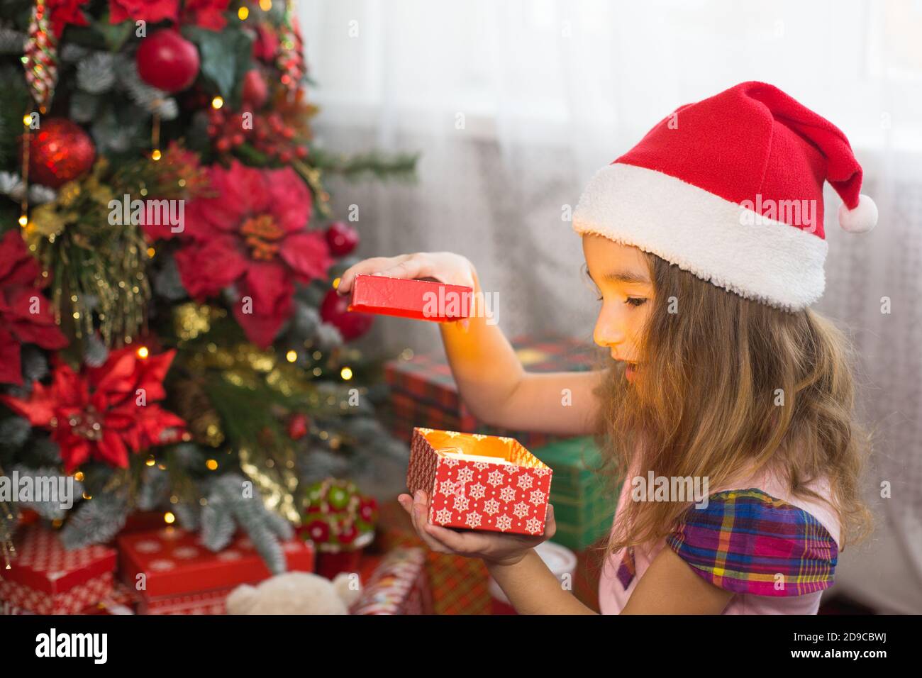Little girl in Santa hat opens a red box with a gift and a Golden magic light near the Christmas tree. Holiday decor, poinsettias on fir trees, New ye Stock Photo