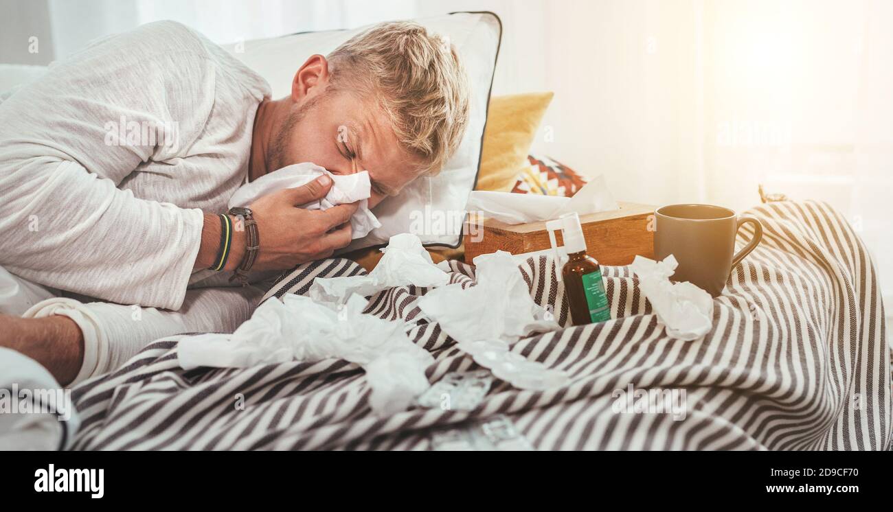 Unhealthy and tired man sneezing or wiping snotty nose man lying in a cozy home bed beside a lot of used paper tissues and medicines on the blanket Stock Photo