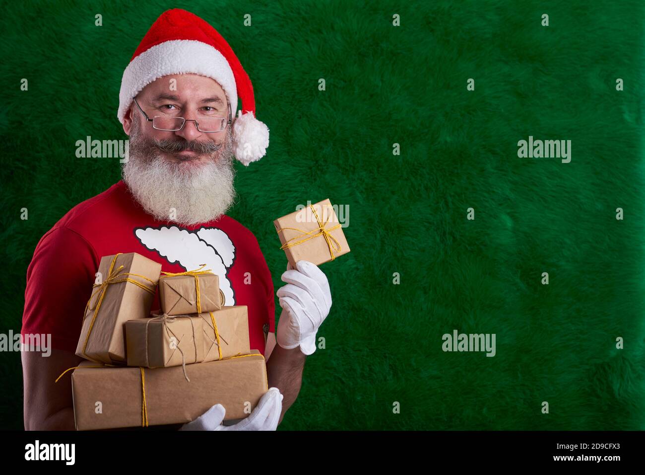 Mature bearded man wearing Santa hat with many gifts in hand, artificial Christmas tree background, copy space Stock Photo