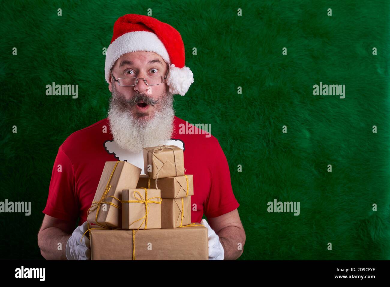 Mature bearded man wearing Santa hat with many gifts in hand, artificial Christmas tree background, copy space Stock Photo