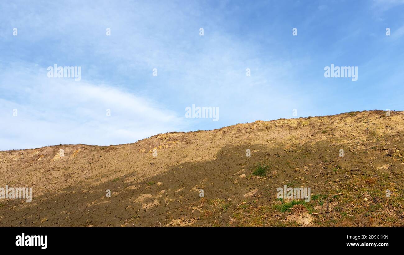 The slope of a clay hill against the backdrop of a beautiful blue sky. Lifeless soil. Sunny weather. Landscape. Stock Photo