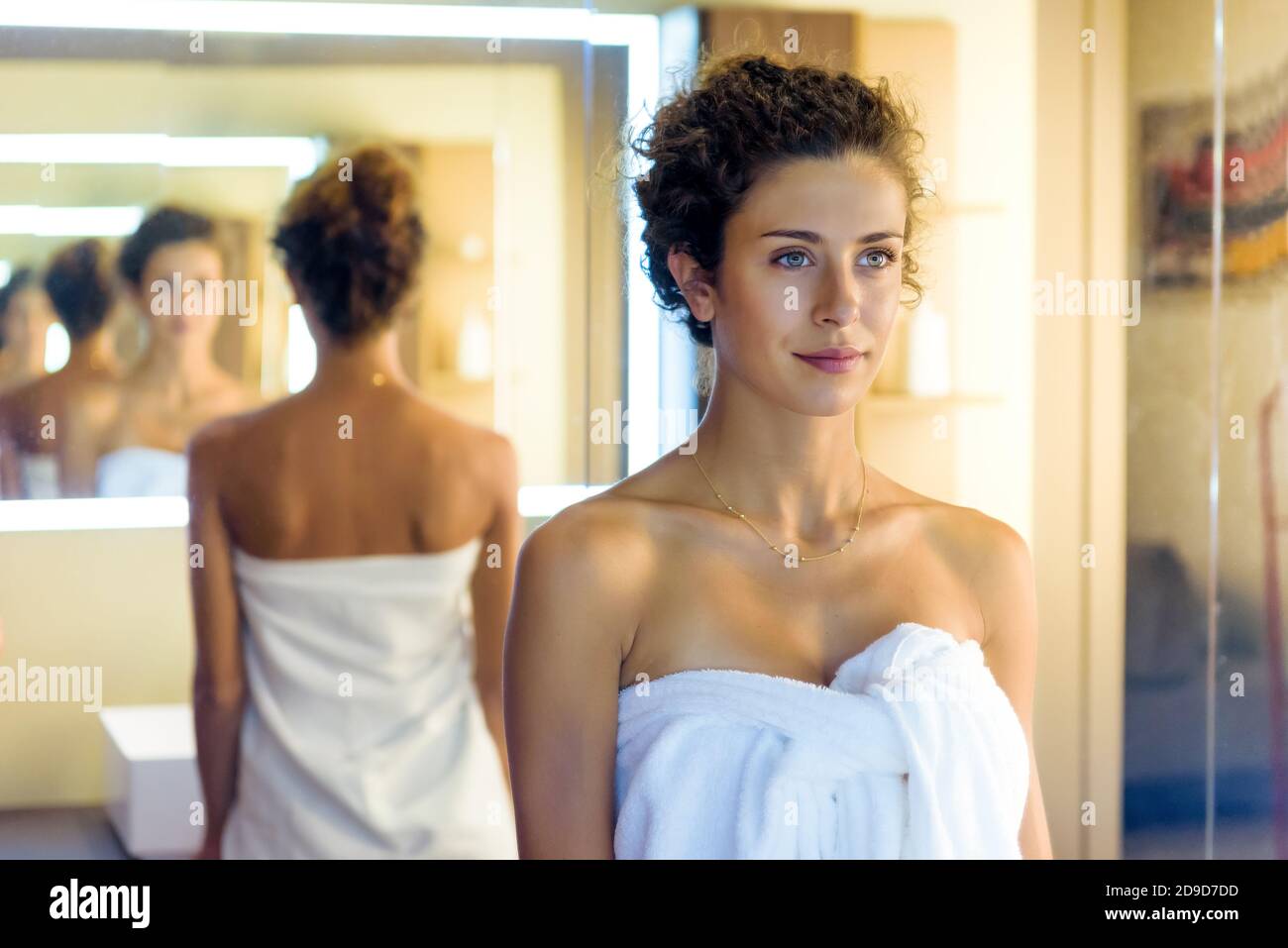 Young woman wrapped in a clean white towel reflecting herself multiple times in parallel mirrors in a diminishing perspective in a a bathroom Stock Photo
