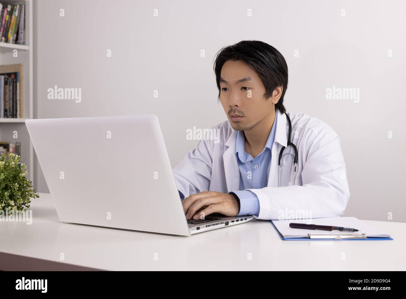 Young Asian Doctor Man in Lab Coat or Gown with Stethoscope Using Laptop Computer on Doctor Table in Office Stock Photo