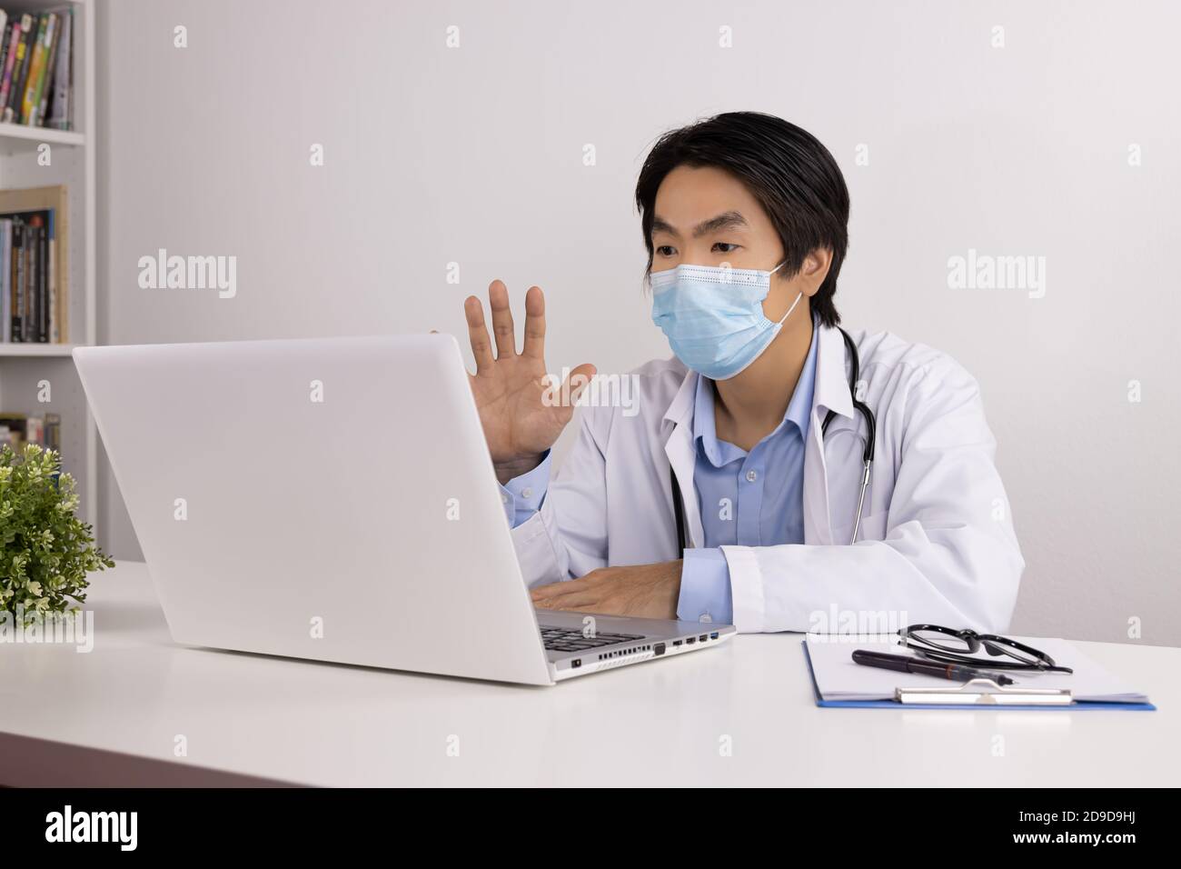 Young Asian Doctor Man in Lab Coat or Gown with Stethoscope Wear Face Mask Video Chat or Video Conference and Talking with Patient Via Laptop Computer Stock Photo