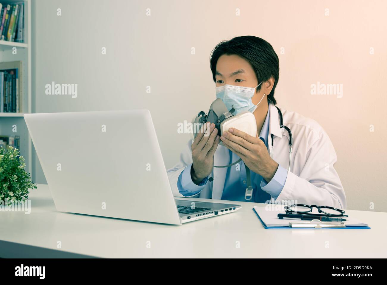 Young Asian Doctor Man in Lab Coat or Gown with Stethoscope Wear Face Mask Demonstrate Filter Mask Using with Patient Via Laptop Computer in Office in Stock Photo