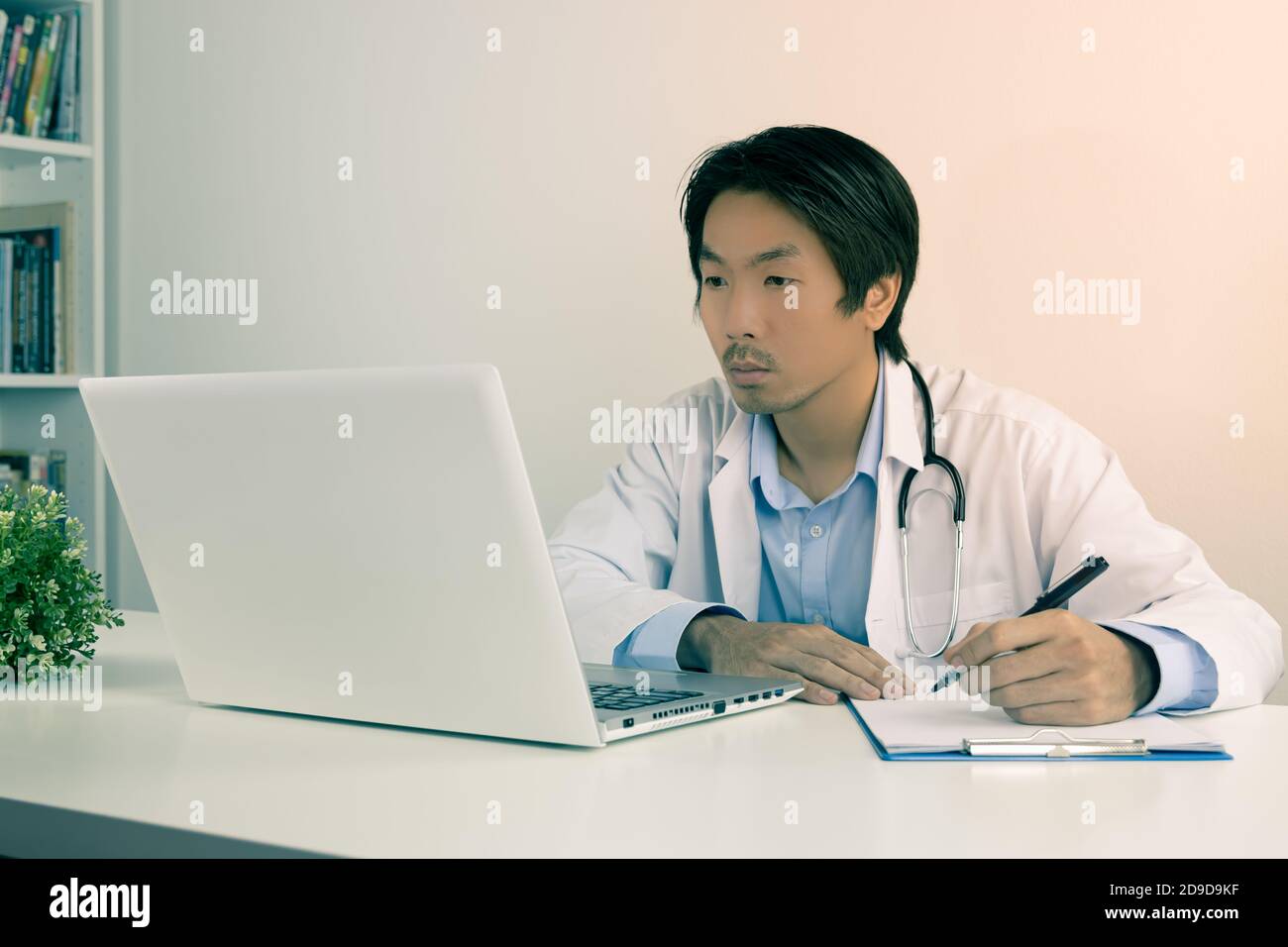 Young Asian Doctor Man in Lab Coat or Gown with Stethoscope Writing Experimental Results Report and Using Laptop Computer on Doctor Table in Office in Stock Photo