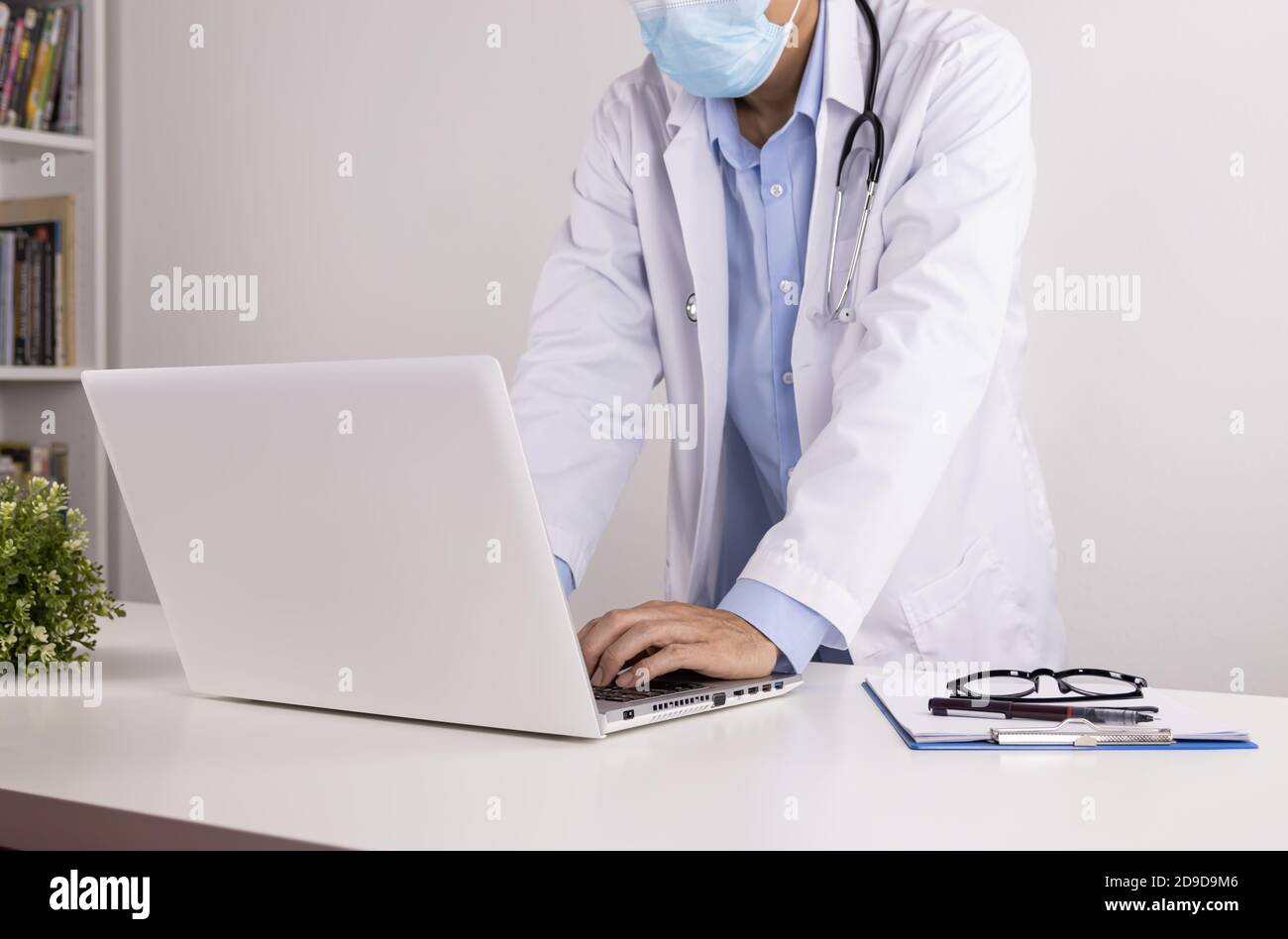Young Asian Doctor Man in Lab Coat or Gown with Stethoscope in Stand Pose Wear Face Mask Using Laptop Computer on Doctor Table in Office Stock Photo