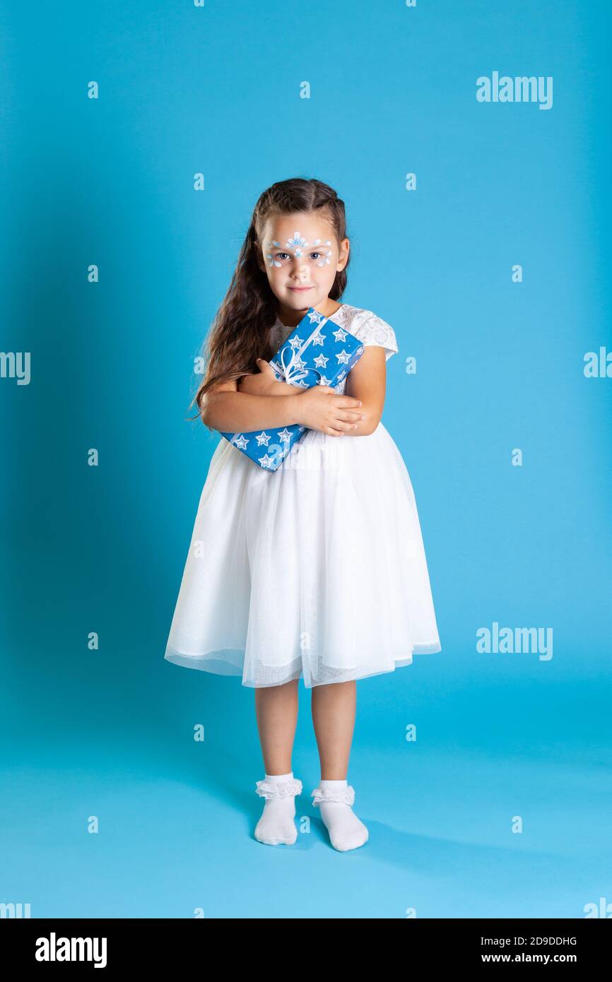 full-length little Princess in white dress and snow petal makeup clutches Christmas gift to her chest, isolated on a blue background Stock Photo
