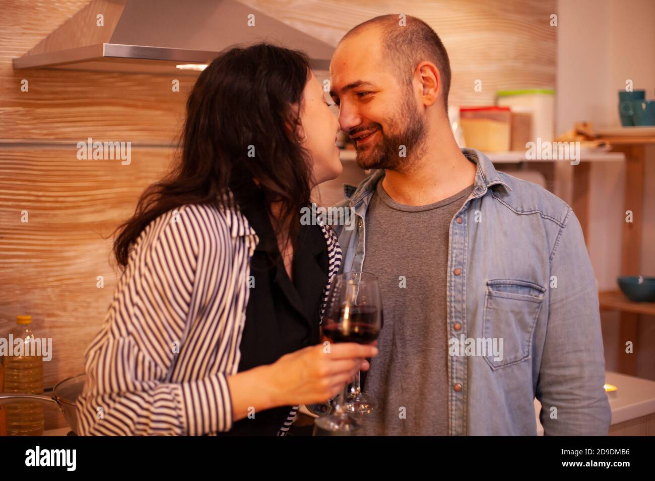 Couple touching noses and having romance date in kitchen. Adult couple at home, drinking red wine, talking, smiling, enjoying the meal in dining room. Stock Photo