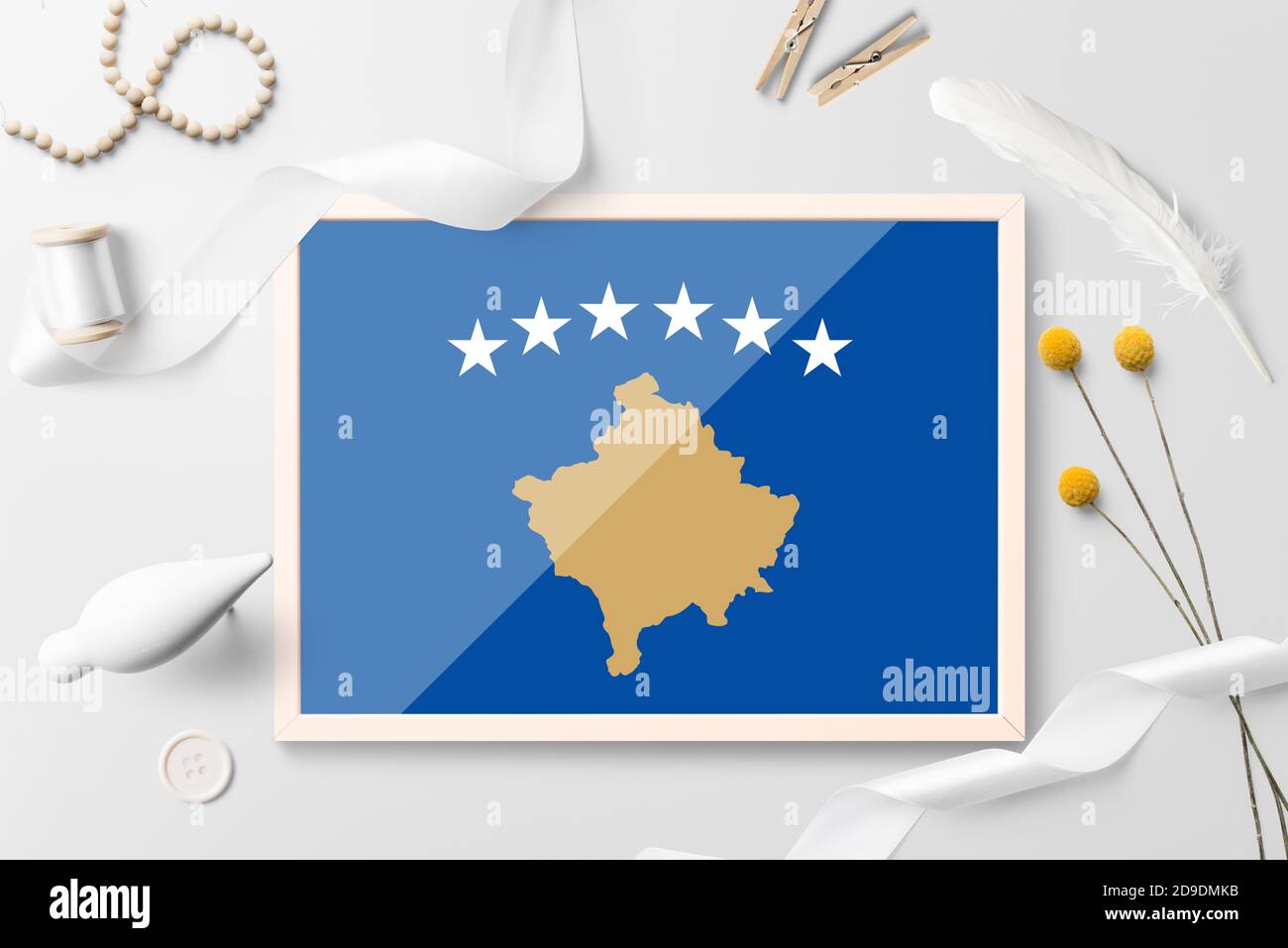 Kosovo flag in wooden frame on white creative background. White theme, feather, daisy, button, ribbon objects. Stock Photo