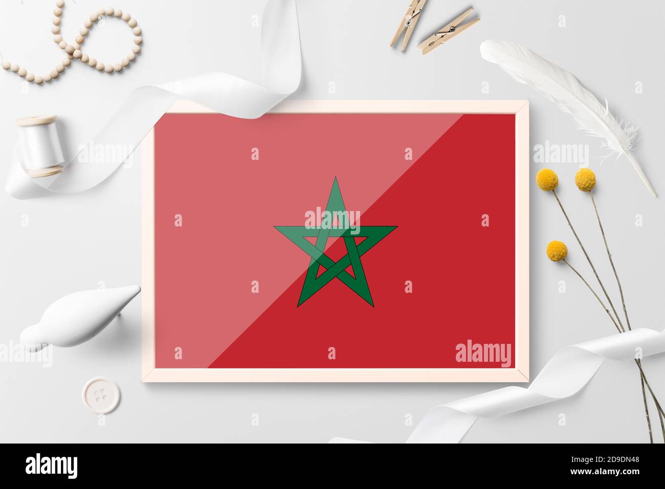Morocco flag in wooden frame on white creative background. White theme, feather, daisy, button, ribbon objects. Stock Photo
