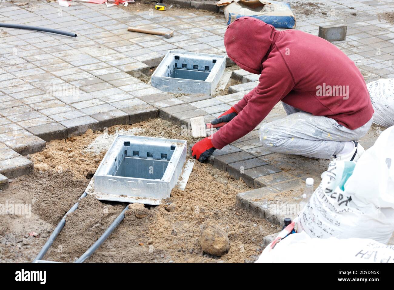 A worker installs engineering hatch shafts and lays paving slabs on prepared flat sandy soil on the sidewalk, copy space. Stock Photo