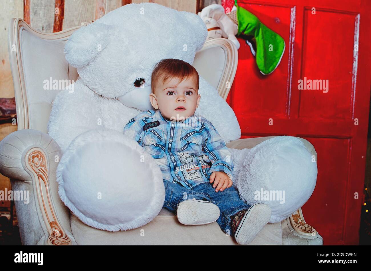 Cheerful and happy little boy sitting with his bear toy on the chair against Christmas decorations. Christmas traditions. Stock Photo