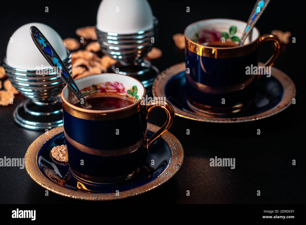 Gourmet breakfast. Two antique enamel cobalt gold rimmed cups of coffee and two boiled eggs on a silver stand on a black background Stock Photo