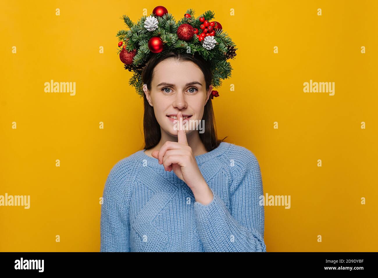 Cute young female saying hush be quiet with finger on lips shhh gesture, wears handmade wreath and blue sweater, isolated on yellow wall studio Stock Photo