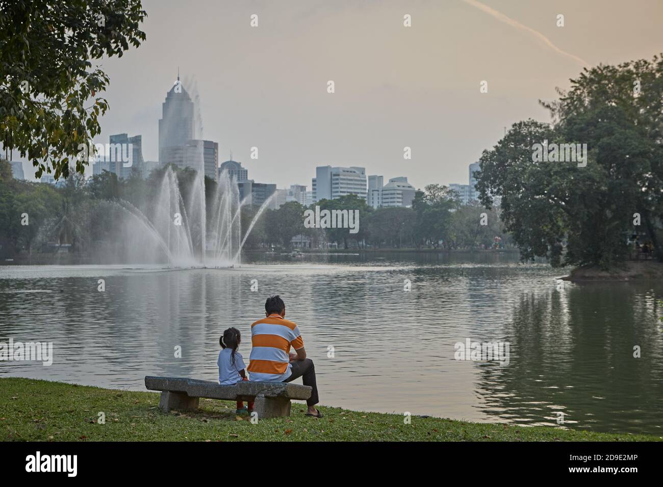 Bangkok, Thailand, March 2106. A father and his daughter sitting on a bench look out over the lake in Lumpini Park. Stock Photo