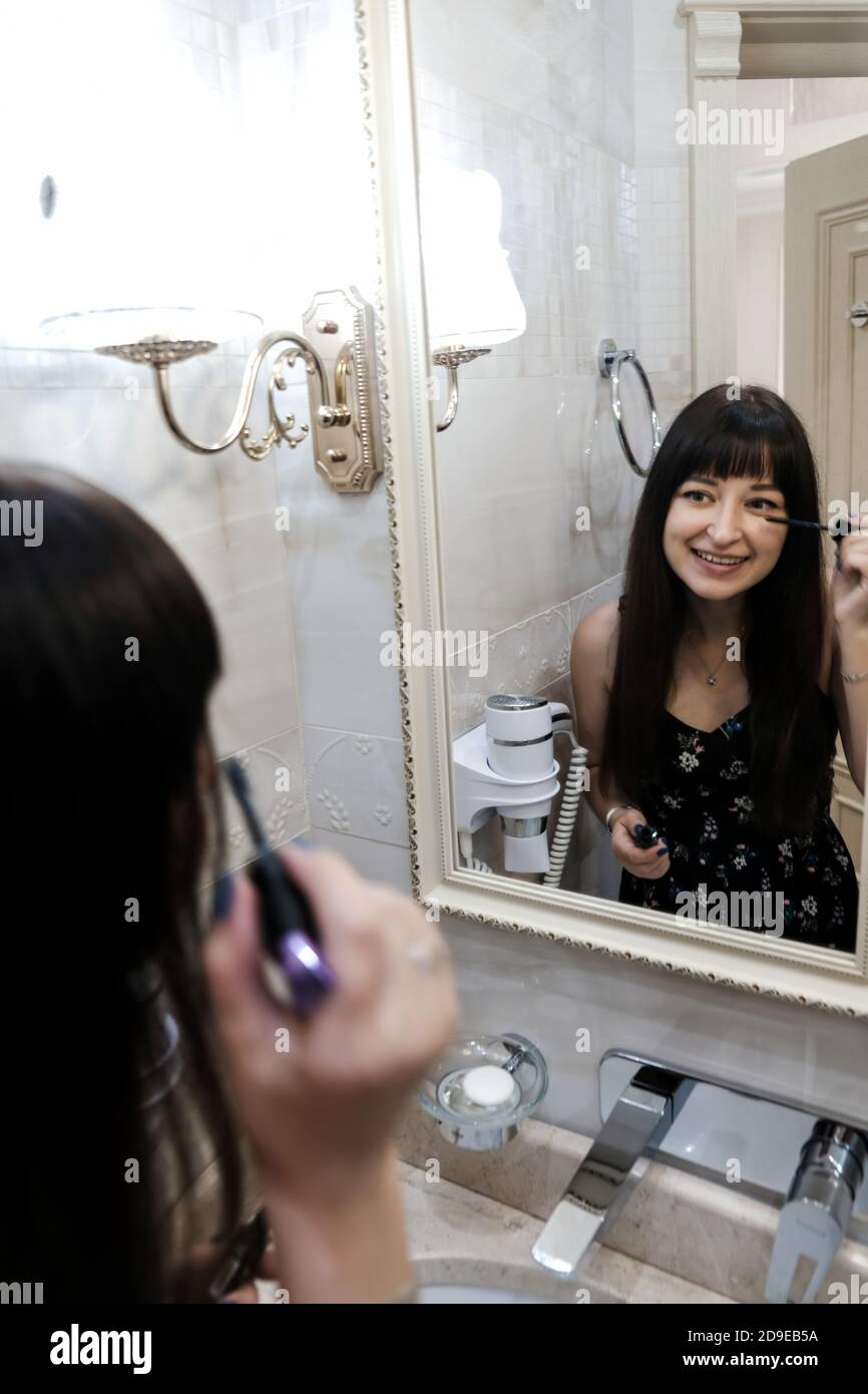 Young woman applying mascara in front of a mirror. Girl getting ready with her makeup in a hotel bathroom. Pretty female smiling while brushing lashes Stock Photo
