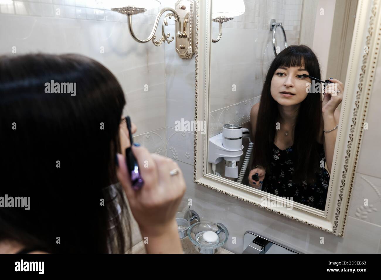 Young woman applying mascara in front of a mirror. Girl getting ready with her makeup in a hotel bathroom. Pretty female serious face while brushing Stock Photo
