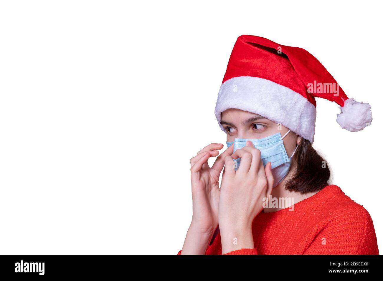 Young teenage girl in red Christmas Santa Claus hat puts on a medical mask, isolated on white background Stock Photo