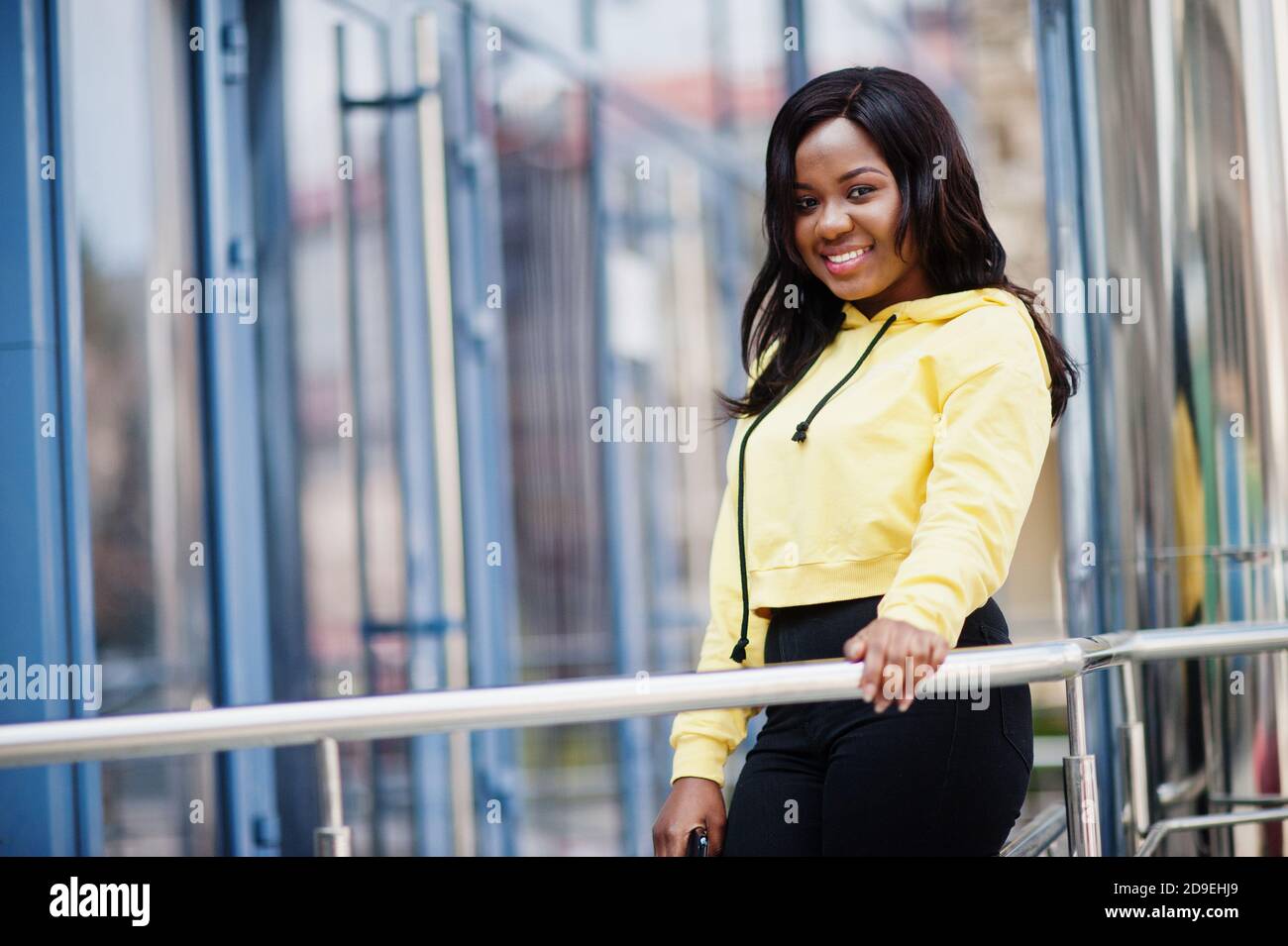 Hipster african american girl wearing yellow hoodie posing at street  against office building with blue windows Stock Photo - Alamy