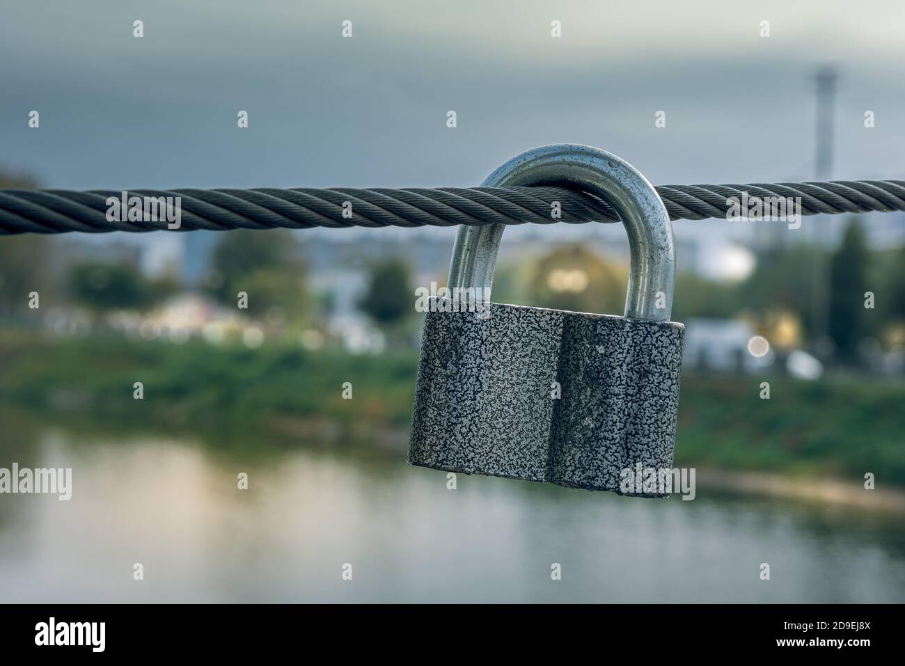 Metal padlock hanging on steel cable of bridge fence. Unfocused cityscape with evening lights at background. Horizontal orientation. Selective focus. Stock Photo