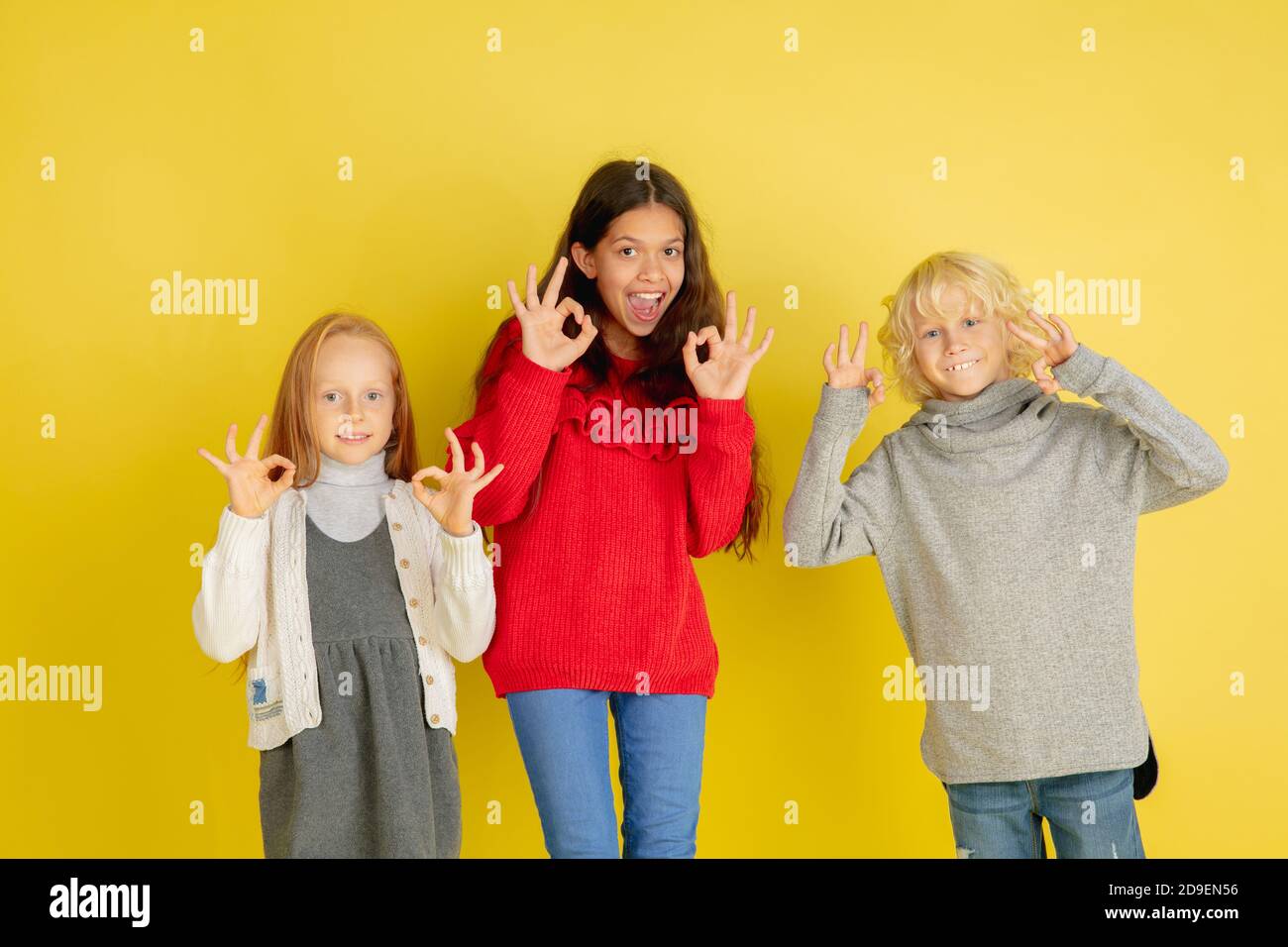 Nice signs, gesturing. Portrait of little caucasian children isolated on yellow studio background with copyspace. Beautiful teen models. Concept of human emotions, facial expression, sales, ad, youth. Stock Photo