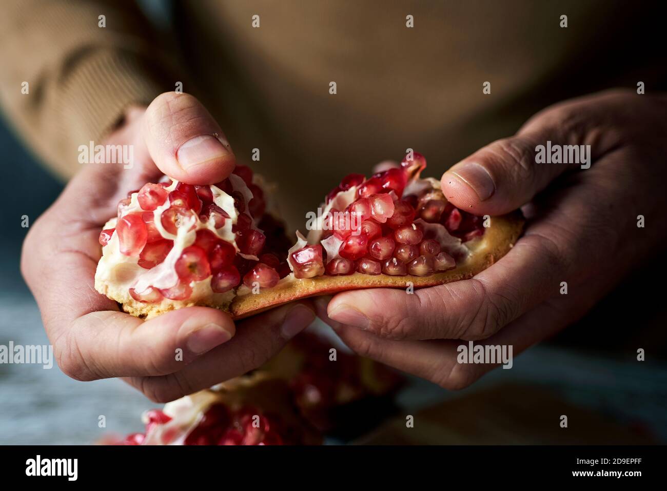 closeup of a young caucasian man, wearing a brown pullover, opening a pomegranate fruit on a rustic wooden table Stock Photo