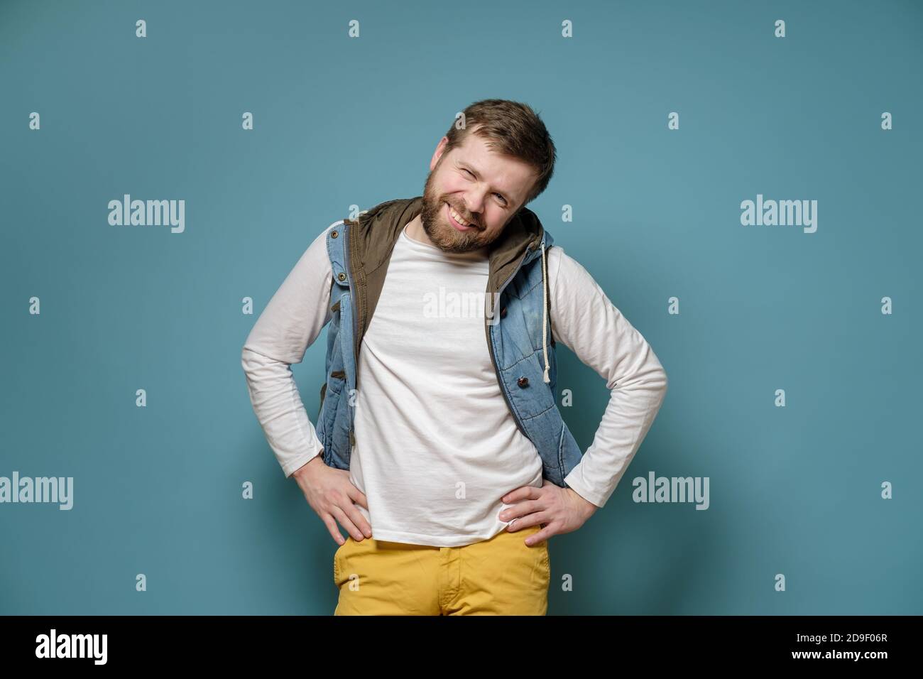 Shy bearded Caucasian man in a vest smiling cute and looking at the camera, on a blue background. Stock Photo