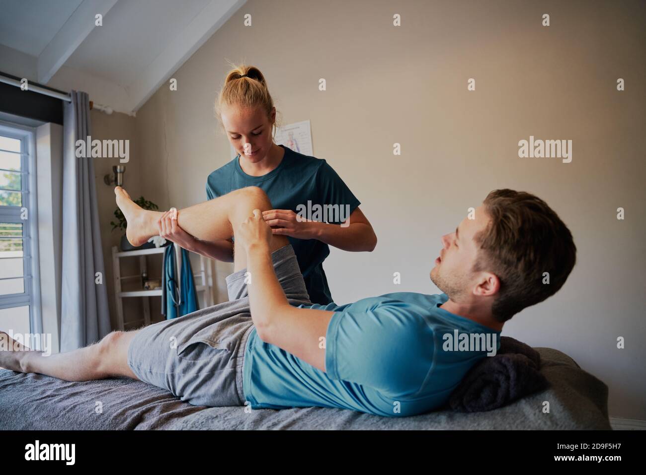 Young man lying on hospital bed showing joint pain position to young woman physiotherapist Stock Photo