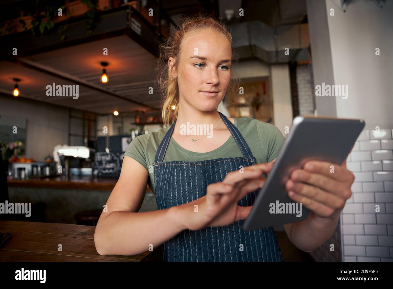 Young female waitress in cafe wearing apron using digital tablet to check online orders Stock Photo