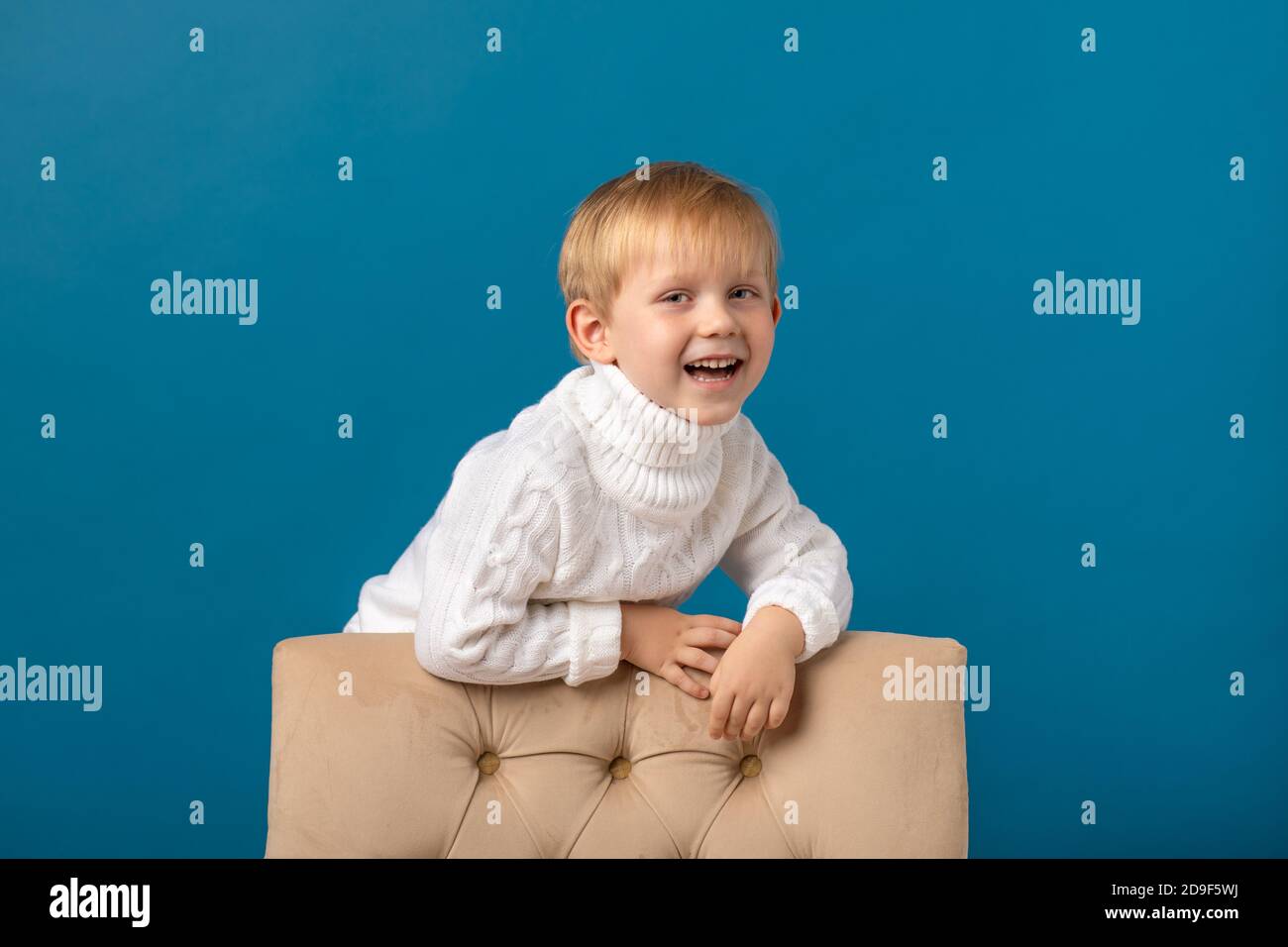A joyful boy, a blond child in a white warm knitted sweater, stands near the back of a chair, looks at the camera and laughs. Winter studio shot on a Stock Photo