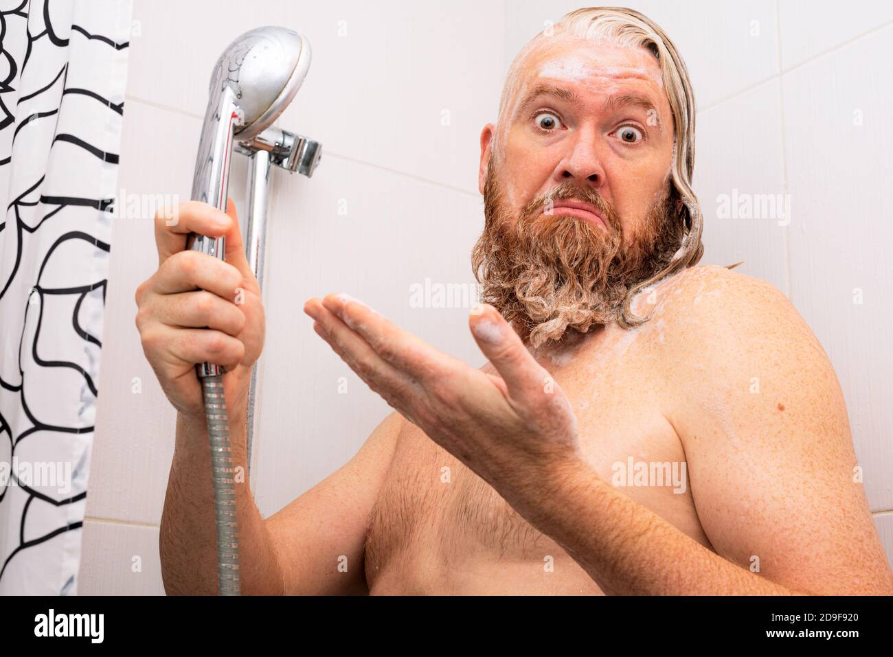 Puzzled wet soapy bearded man standing in bathtub holding shower in his