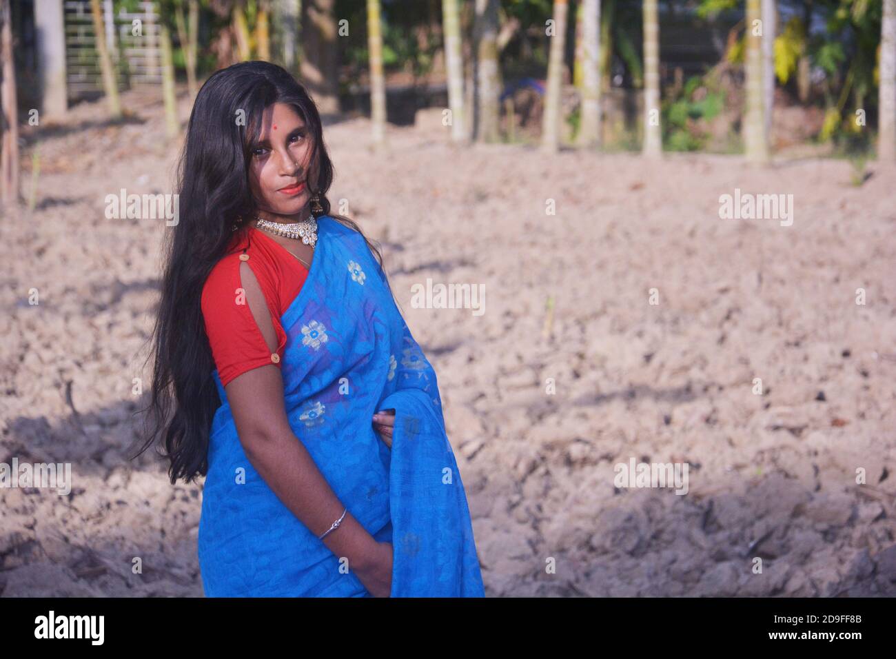 Close up side face of an Indian teenage girl wearing blue sari with red blouse nose pin big earring standing on a plowed field, selective focusing Stock Photo
