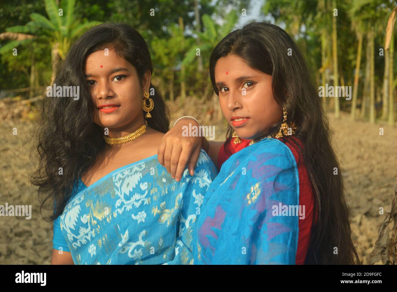 Two beautiful Indian Bengali Teenage girls wearing blue sari, necklace earrings nose pin posing in a plowed agricultural field one hand on shoulder. Stock Photo