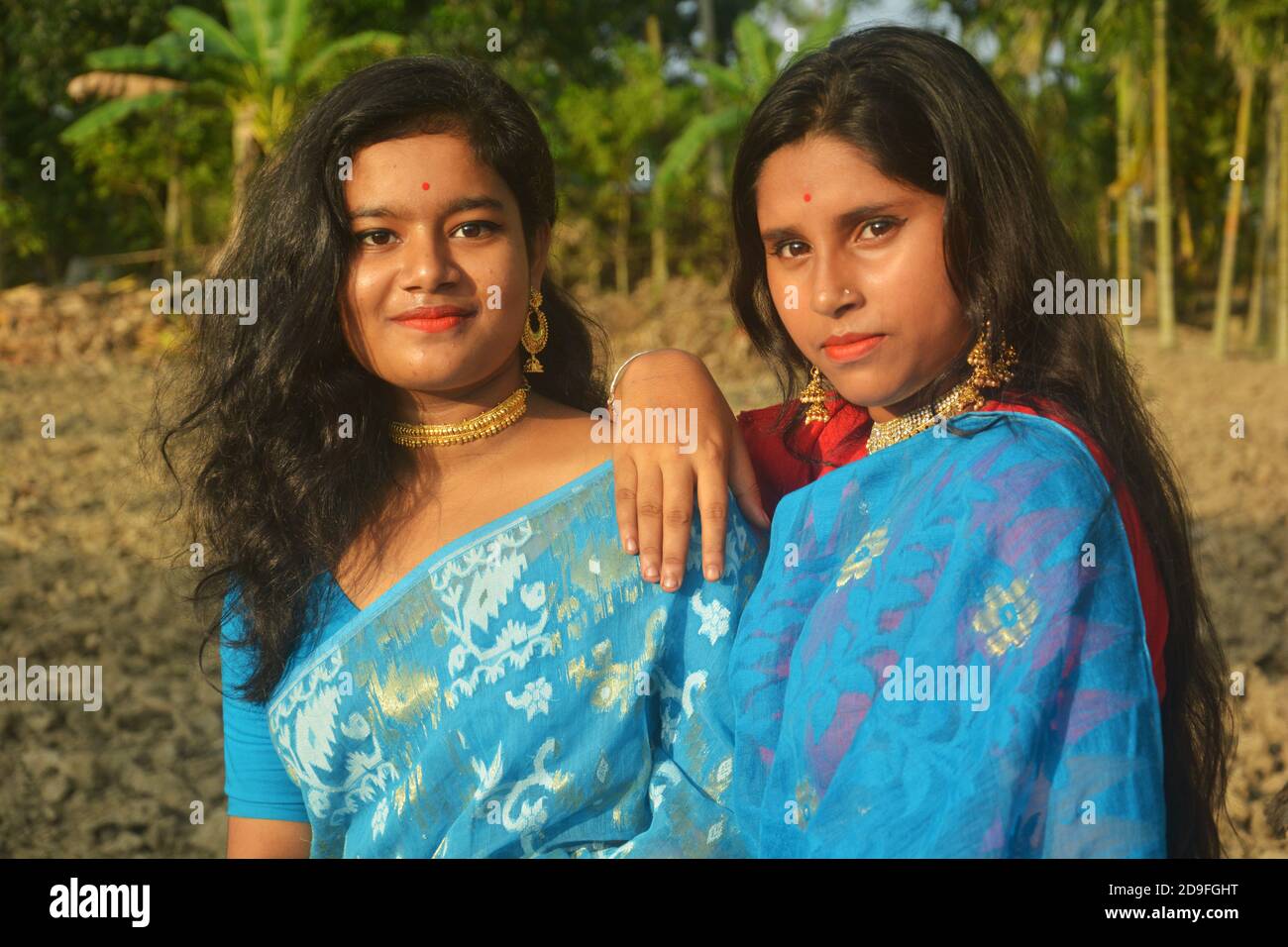 Two beautiful Indian Bengali Teenage girls wearing blue sari, necklace earrings nose pin posing in a plowed agricultural field one hand on shoulder. Stock Photo