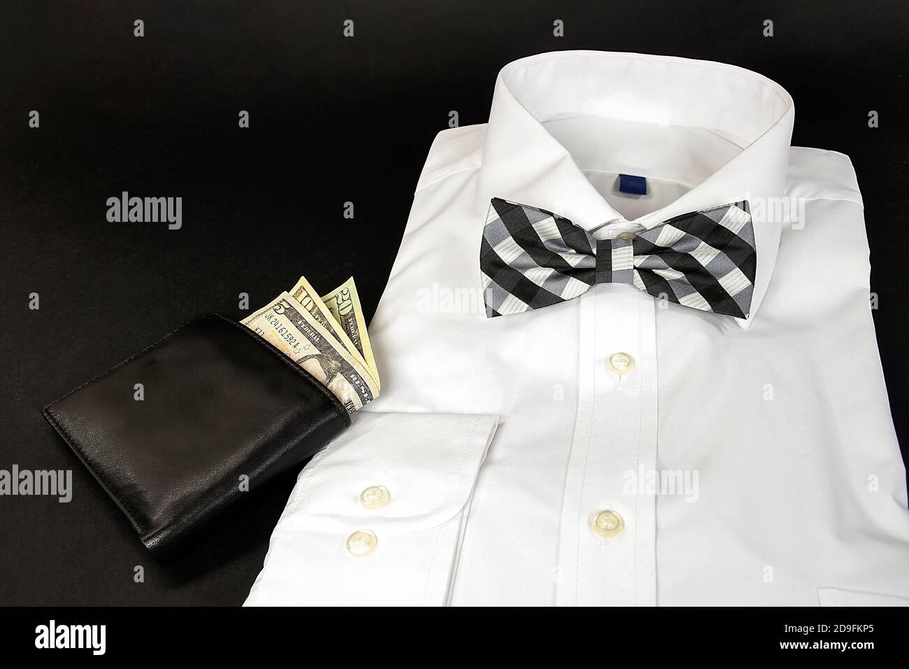 Folded white dress shirt with black and white plaid bow tie and leather wallet on black background Stock Photo