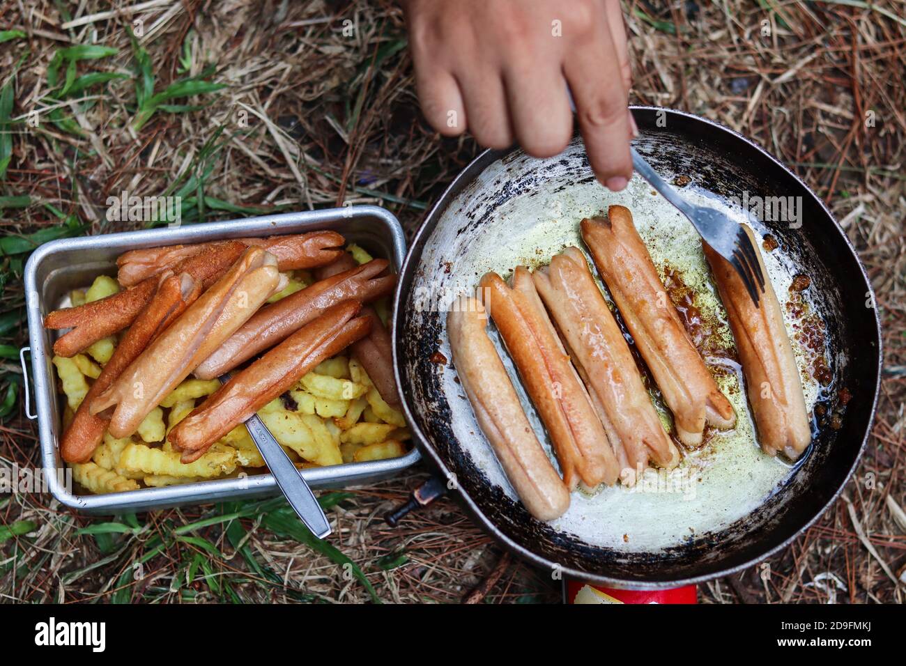 hand frying potatoes and chicken sausages in the outdoor on a red mini gas stove and black frying pan Stock Photo