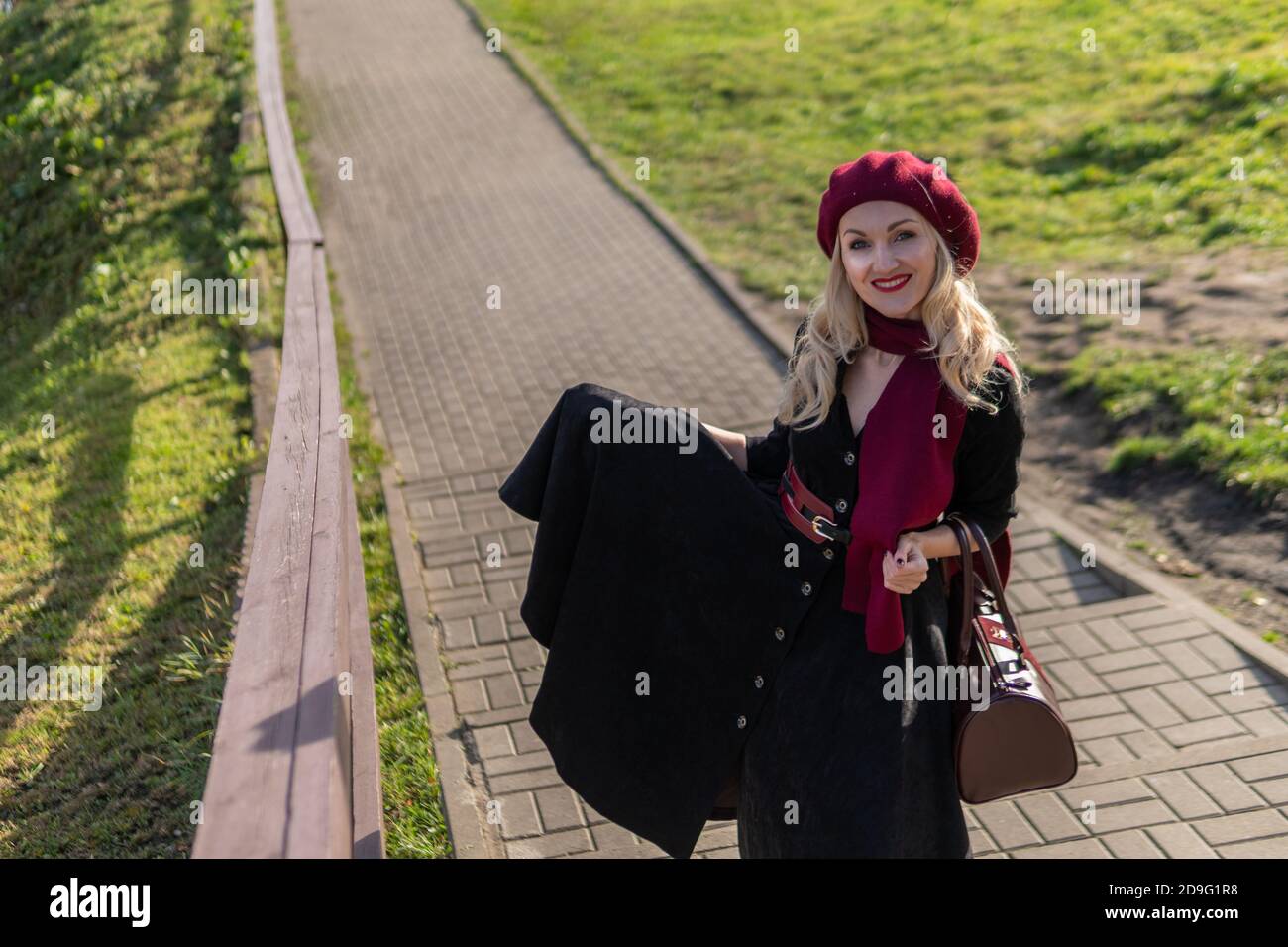a girl walks up the stairs in a burgundy palette and biret, in black clothes, in the fall against the background of a pond there are blue clouds. Stock Photo